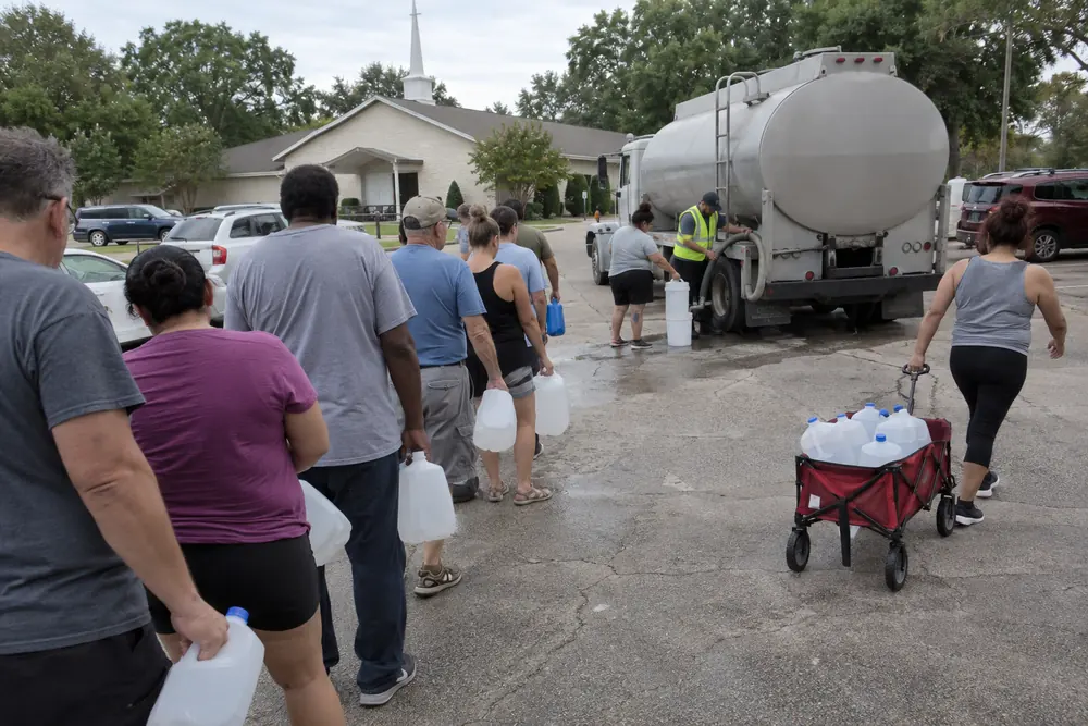 Residents in a long line with empty jugs and buckets at an emergency water tanker in a parking lot