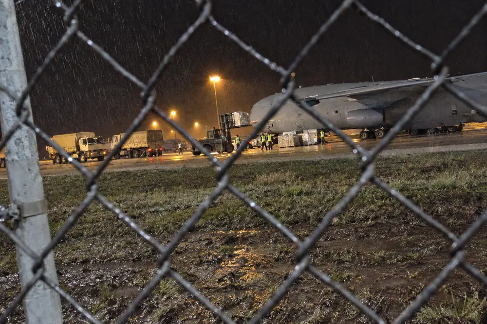 Airmen loading pallets of gear and generators into a military cargo plane under floodlights at night, seen through the air base fence