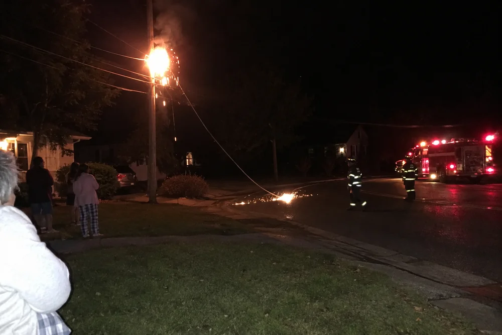 A blown transformer burning on top of a utility pole at night, a line down in the street and a fire engine stopped short, neighbours watching