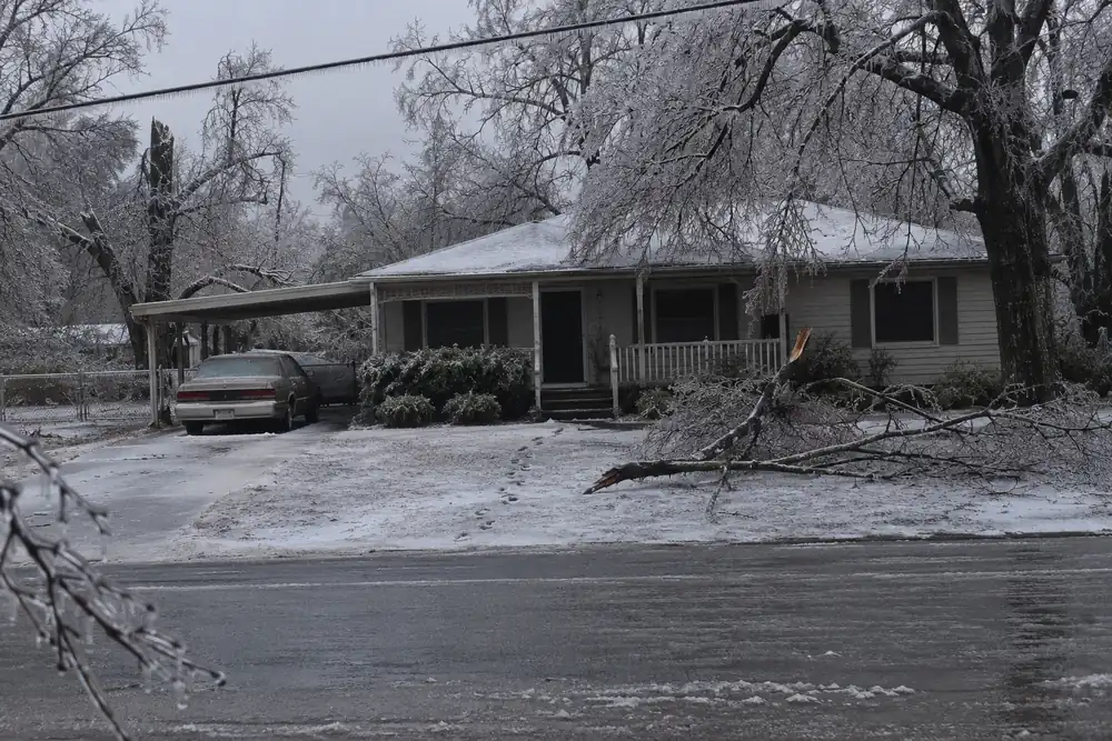 A small one-story house in the rural South at dusk after an ice storm, every window dark, ice on the trees, the porch rail and the line, a car in the drive glazed with ice