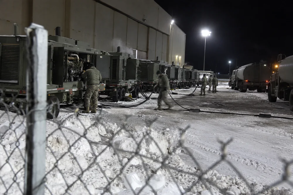 A military air base on a winter night seen through the perimeter fence: a row of trailer generators and fuel trucks under floodlights, airmen in parkas running cables in the snow