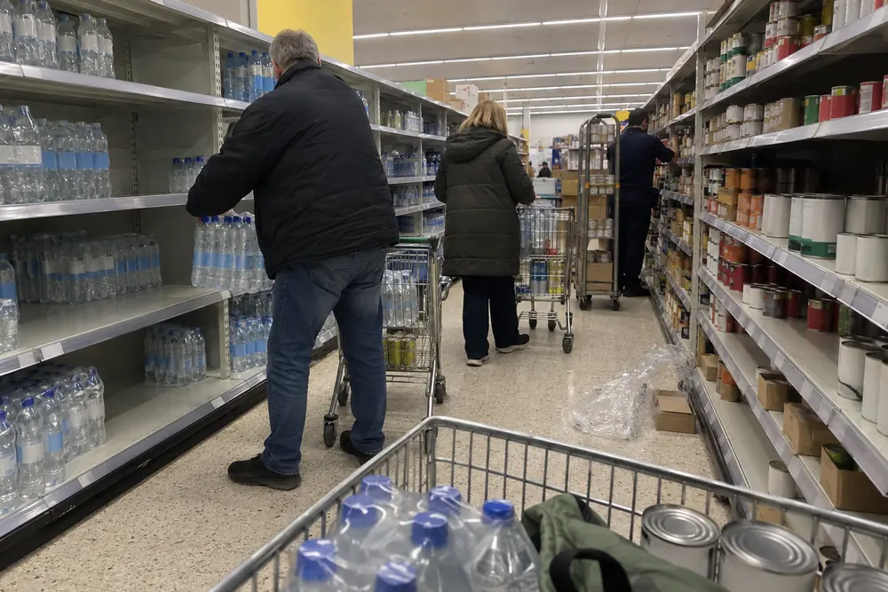 Shoppers in coats loading bottled water and tinned food into trolleys in a half-empty British supermarket aisle
