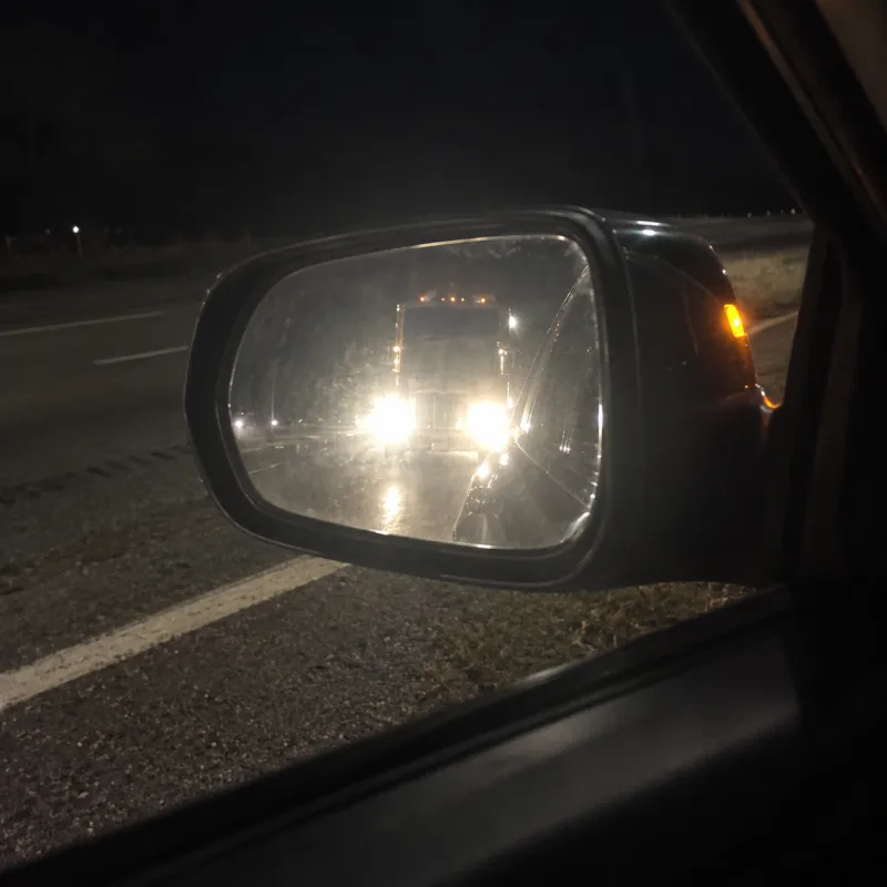 Seen from her driver's seat at night, the side mirror is full of a semi truck's blinding headlights.