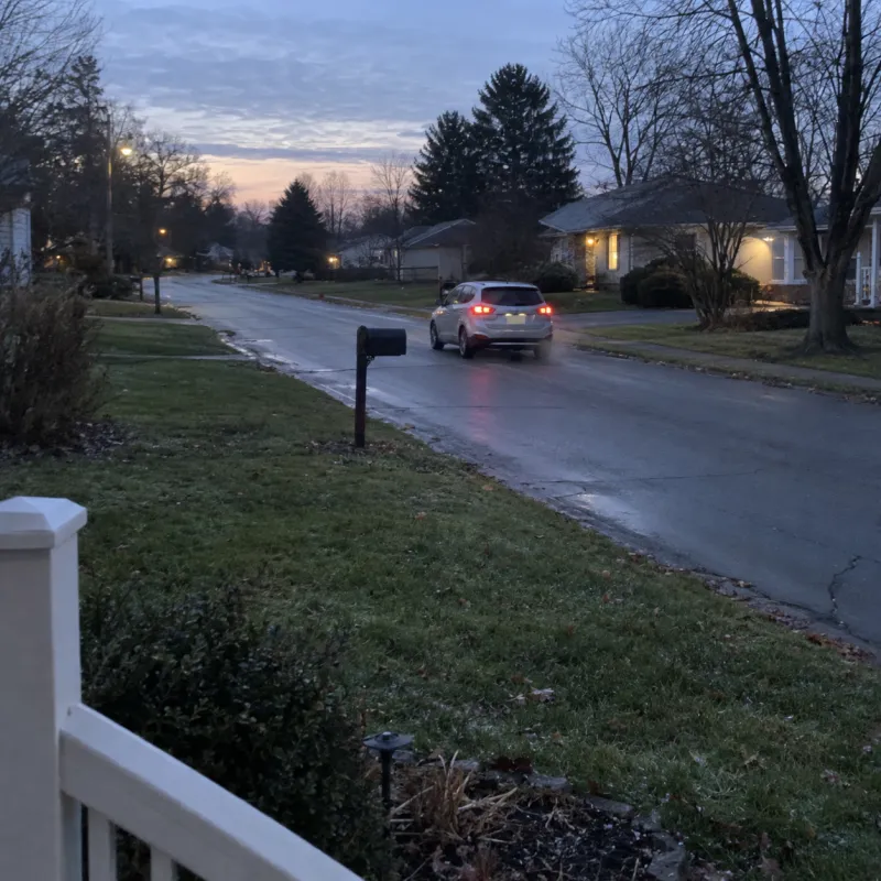 At dawn, her grandson's small silver car pulls away down a quiet street on his first drive alone, seen from her front porch.