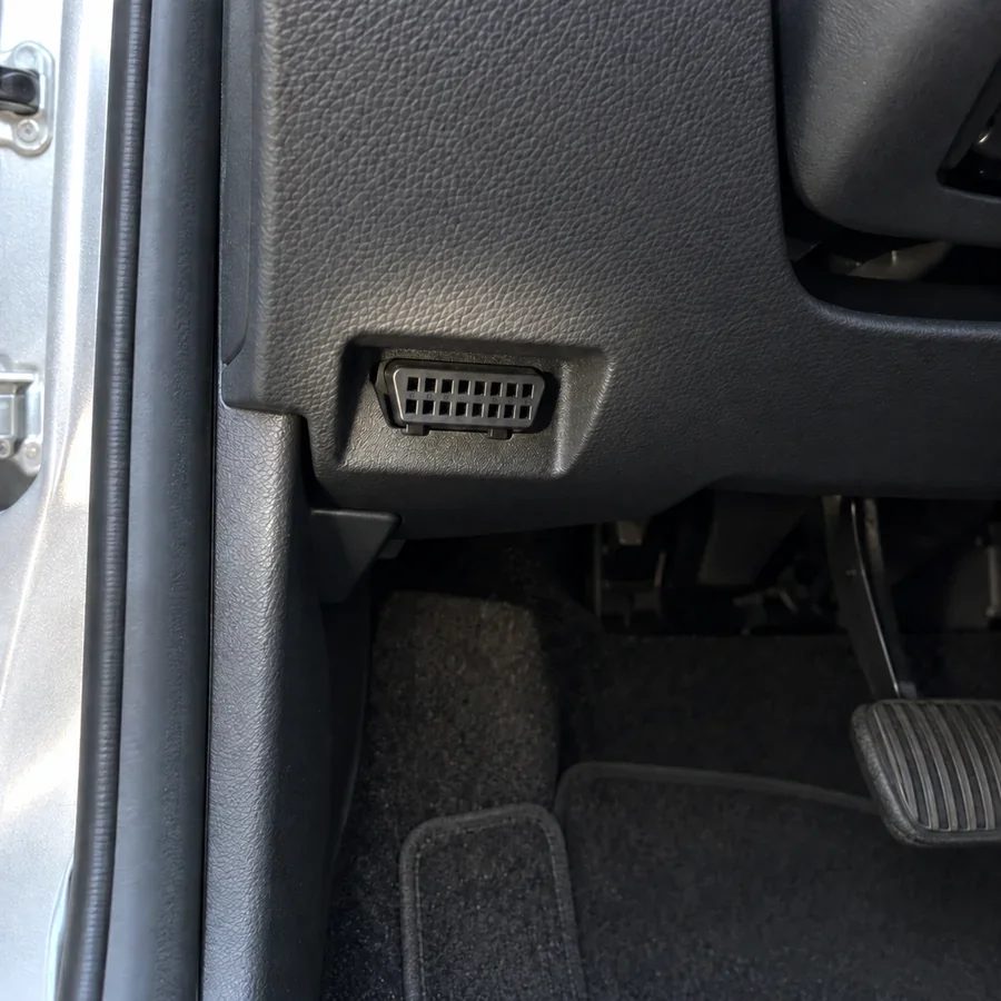 The empty socket with 16 small holes under a car's dash, near the driver's left knee, lit by a flashlight.