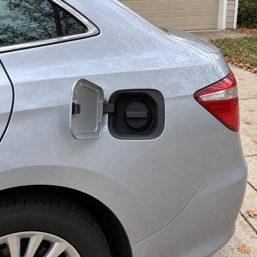 The open fuel door of her silver car in the driveway, the black gas cap screwed back in tight.