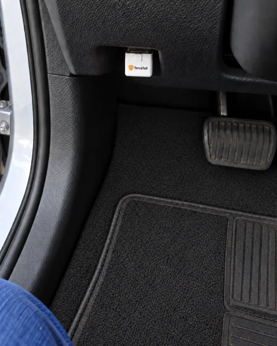Looking down past her left knee: the small white TerraTell plugged into the socket under the dash, above the brake pedal.